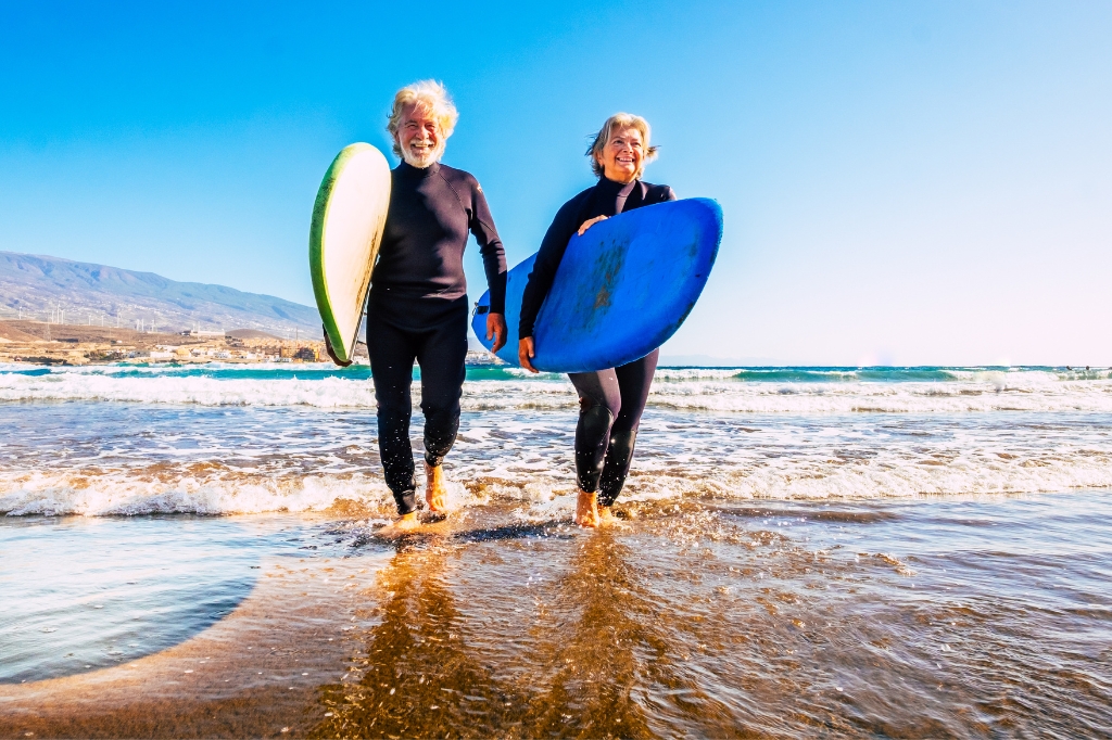 An elderly couple walks along the beach with surfboards